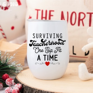 May include: White wine tumbler with black text that reads "Surviving Teacherhood One Sip At A Time" with a red heart in the center.