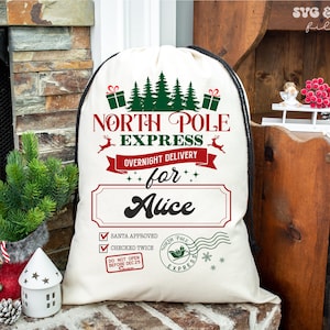 May include: A cream-coloured drawstring sack featuring a North Pole Express graphic in red and green. The design includes pine trees, gift boxes, reindeer, and a personalised name field with the name Alice. It also features Santa approved and checked twice checkboxes, a North Pole Express postmark, and a do not open before Dec 25 stamp.