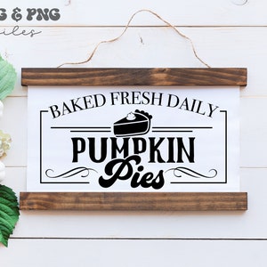 May include: A rectangular wall hanging sign with dark brown wooden slats at the top and bottom, featuring a white center panel with black text that reads BAKED FRESH DAILY PUMPKIN Pies and a graphic illustration of a slice of pumpkin pie.
