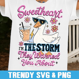 Könnte beinhalten: Weißes T-Shirt mit einer Grafik einer Comic-Gans mit Sonnenbrille und Eiskaffee. Der Text lautet: "Sweetheart, I'm the storm they warned you about." Rosa und blaue Akzente.