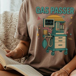 Puede incluir: Camiseta marrón con el texto "GAS PASSER" sobre una ilustración retro de un respirador médico. El respirador es de color turquesa, amarillo y rosa con un monitor que muestra un latido cardíaco. La camiseta tiene las mangas dobladas.