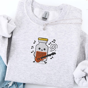 Może przedstawiać: Jasnoszara bluza z nadrukiem z kreskówki przedstawiającym fiolkę grającą na gitarze. Fiolka ma uśmiechniętą twarz i napis "Roc-unium" na etykiecie. Nuty otaczają fiolkę. Srebrny naszyjnik i dżinsy są również na zdjęciu.