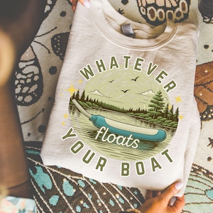 May include: A light beige t-shirt with a circular graphic featuring a canoe on a lake, trees, and the text "Whatever Floats Your Boat." The design includes a blue canoe and green trees. The shirt is folded.