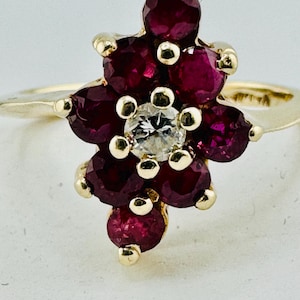 May include: A gold ring featuring a cluster of deep red gemstones arranged in a floral pattern, surrounding a central clear gemstone. The ring's band is a simple, polished gold.