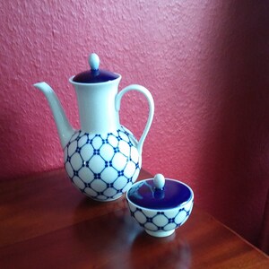 Pode incluir: Um conjunto de chá de porcelana branca com um padrão geométrico azul. O conjunto inclui uma chaleira com tampa e um açucareiro pequeno com tampa. A chaleira tem um corpo arredondado e um bico curvo. O conjunto é exibido em uma superfície de madeira.