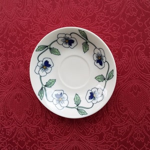 Könnte beinhalten: Weiße Keramikuntertasse mit einem Blumendesign mit blauen und weißen Stiefmütterchen und grünen Blättern. Die Untertasse steht vor einem roten, gemusterten Hintergrund. Das florale Muster umrandet den Rand und bietet eine zarte Ästhetik.