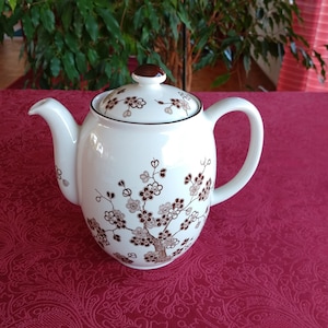 Könnte beinhalten: Weiße Keramik-Teekanne mit braunem Blumendesign. Die Teekanne hat einen gebogenen Ausguss, einen abgerundeten Körper und einen passenden Deckel mit einem braunen Knauf. Das Blumenmuster umfasst braune und hellbraune Blüten und Zweige. Die Teekanne steht auf einer roten Tischdecke.