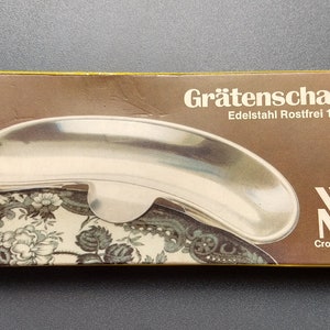 Könnte beinhalten: Eine Grätenschale aus Edelstahl in der Originalverpackung. Die Schachtel ist braun mit dem Text "Grätenschale Edelstahl Rostfrei 18/10" und dem WMF Cromargan Logo. Die Schale ist silberfarben und hat eine einzigartige, geschwungene Form.