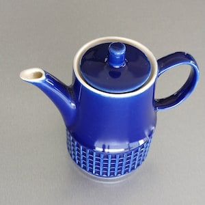 Könnte beinhalten: Eine kobaltblaue Keramik-Teekanne mit passendem Deckel und Griff. Die Teekanne hat ein strukturiertes, gitterartiges Muster um den Boden und einen weißen Rand. Der Ausguss ist weiß. Ein klassisches Design zum Servieren von Tee oder Kaffee.