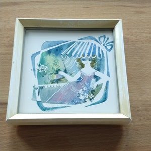 Könnte beinhalten: Quadratisch gerahmtes Kunstwerk mit einer sitzenden Frau unter einem blau-weiß gestreiften Sonnenschirm. Das Kunstwerk hat einen Aquarellstil mit Blau-, Grün- und Lilatönen. Der Rahmen ist hellbeige.