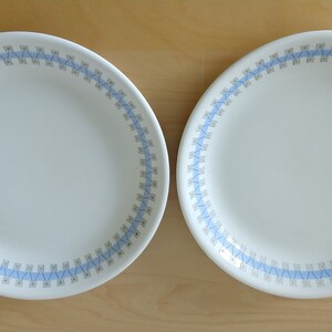 May include: Two white ceramic plates with a light blue and gray geometric border design. The plates are round and have a simple, elegant aesthetic, suitable for everyday use or special occasions.