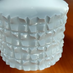 Puede incluir: Un recipiente de cerámica blanca con un diseño geométrico texturizado. El cuerpo cilíndrico presenta un patrón de formas cuadradas en relieve. La parte superior es ligeramente acampanada y la superficie tiene un acabado brillante que refleja la luz.