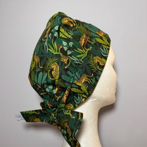 Peut inclure: Un bonnet de chirurgien vert foncé avec un motif sur le thème de la jungle. Le tissu présente des léopards jaunes, des feuilles vertes et du feuillage. Le bonnet est noué dans le dos avec un nœud.