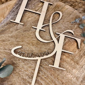Könnte beinhalten: Ein Kuchenaufsatz aus Holz mit den Buchstaben "H & F" in einer stilisierten Schriftart und dem Datum "27.06.2026" darunter ausgeschnitten. Der Aufsatz ist aus hellem Holz gefertigt und für eine Hochzeits- oder Jubiläumsfeier gedacht.