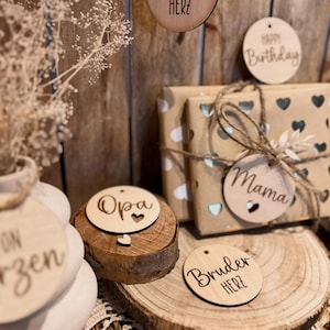 Könnte beinhalten: Holz-Geschenk-Etiketten mit verschiedenen Botschaften, darunter "Schwester Herz", "Happy Birthday", "Opa", "Mama" und "Bruder Herz". Die Etiketten befinden sich auf einer Holzoberfläche mit braunem Hintergrund.