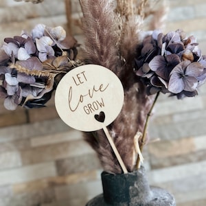 Könnte beinhalten: Ein Holzschild mit den Worten "LET LOVE GROW" und einem Herz-Ausschnitt, platziert in einer Vase mit getrockneten Blumen und Pampasgras. Die Vase ist dunkelgrau und hat eine strukturierte Oberfläche. Die Blumen sind eine Mischung aus Lila und Braun.