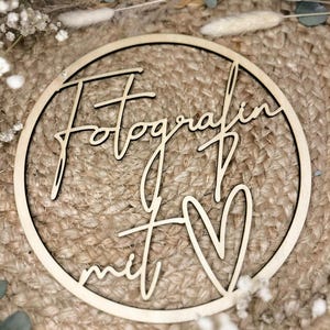 Könnte beinhalten: Runder Holzschild mit den Worten "Fotografin mit" und einem ausgeschnittenen Herz. Das Schild ist hellbraun mit schwarzem Umriss und liegt auf einer gewebten Oberfläche, umgeben von Trockenblumen.