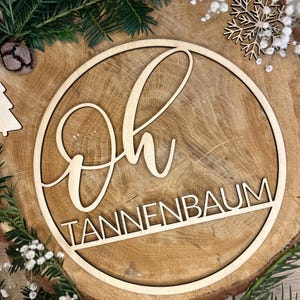 Könnte beinhalten: Ein kreisförmiges Holzschild mit den Worten "Oh Tannenbaum" ausgeschnitten. Das Schild ist von Grün und kleinen weißen Blumen umgeben, mit einer Holzschneeflocke und einem Tannenzapfen im Hintergrund. Das Schild ist aus hellem Holz.