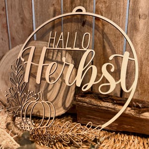 Könnte beinhalten: Ein kreisförmiges Wandobjekt aus Holz mit dem lasergeschnittenen Schriftzug "HALLO Herbst" in einer Kombination aus Block- und Schreibschrift. Das Design zeigt unten links ein Kürbis- und Blattmotiv aus hellem, glattem Holz.