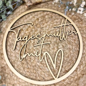 Könnte beinhalten: Ein runder, hellfarbiger Holzschild mit den Worten "Tagesmutter mit" in einer Schreibschrift und einer Herzform. Das Schild ist vor einem strukturierten, gewebten Hintergrund mit Grün und kleinen weißen Blumen im Hintergrund platziert.