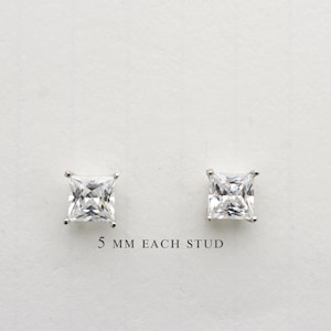 May include: Two square-cut, clear gemstone stud earrings, each measuring 5 mm, are displayed against a white background. The earrings are set in a silver-colored metal, showcasing their brilliant facets. The text "5 mm EACH STUD" is printed below the earrings.