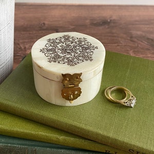 May include: A small, round, white box with a brass latch and intricate floral design on the lid. The box is sitting on a stack of green books.