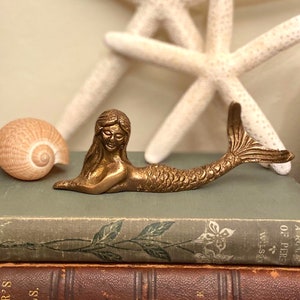 Puede incluir: Una figurita de sirena de latón dorado con una cola larga y una cara detallada. La sirena está tumbada sobre una pila de libros antiguos con una concha a la izquierda.