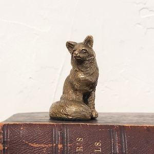 May include: A small, gold-colored metal figurine of a fox sitting on a book. The fox is detailed with a textured surface and has a curled tail.