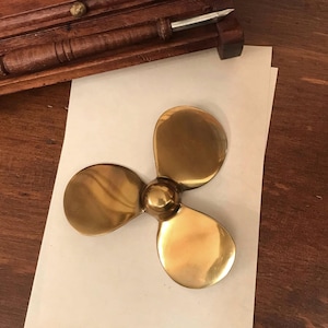 May include: A polished brass three-blade boat propeller with a central hub. The propeller is resting on a piece of off-white paper, positioned below a dark brown wooden pen holder containing a metal-tipped fountain pen.