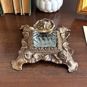 Könnte beinhalten: Ein antikes Tintenfass aus Messing mit einem eulenartigen Deckel. Das Tintenfass hat einen quadratischen Glaseinsatz und ist in einem verzierten, detaillierten Messingrahmen gefasst. Das Stück steht auf einer dunklen Holzoberfläche.