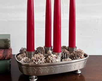 May include: An oval, silver-toned candle holder with four red taper candles. The holder is decorated with pinecones and small, decorative flowers. The holder has a decorative pattern around the edge and rests on four small feet.