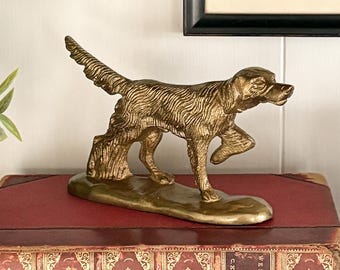 May include: A metallic gold-toned brass figurine of a hunting dog in a standing position with one front paw raised. The dog has a textured coat and is mounted on a solid brass base. The sculpture is captured in a side profile view.