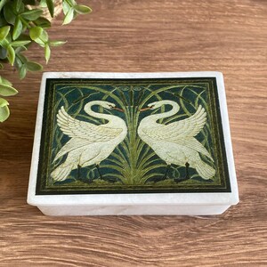 May include: A rectangular white stone box featuring a decorative top panel with two white swans facing each other. The swans are set against a dark green background with stylised green reeds and cattails, framed by a thin black border.