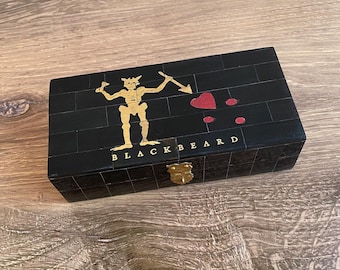 Puede incluir: Una caja de madera negra con un esqueleto dorado sosteniendo una lanza y un corazón. La caja tiene un pestillo dorado y el texto "BLACKBEARD" en la parte delantera.