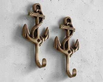 May include: Two identical brass-colored metal wall hooks shaped like nautical anchors. Each hook features a central vertical shank wrapped in a rope design, a horizontal crossbar at the top, two curved flukes at the base, and a single hook extending downward.