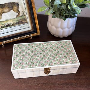 Puede incluir: Caja decorativa rectangular con un estampado floral en la tapa. La caja es blanca con un cierre de latón. La tapa presenta un patrón repetido de flores rosas y hojas verdes sobre un fondo verde claro. La caja está sobre una superficie de madera oscura.