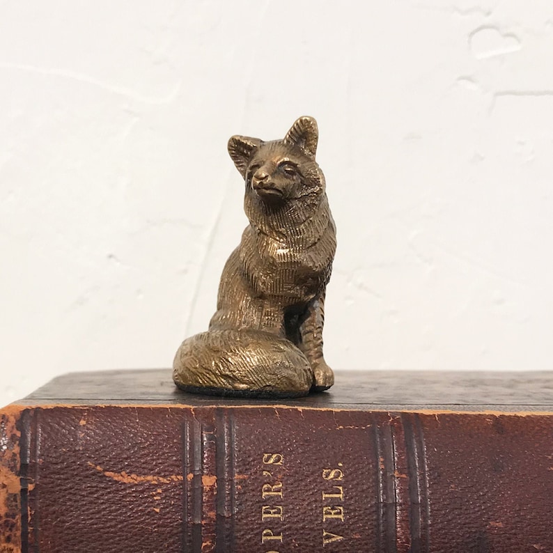May include: A small, gold-colored metal figurine of a fox sitting on a book. The fox is detailed with a textured surface and has a curled tail.