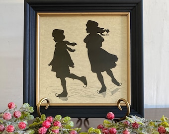 May include: Framed silhouette art featuring two figures ice skating. The black silhouettes are set against a cream-colored background, framed in black with a gold inner border. The artwork is displayed on a dark surface with a decorative berry garland.