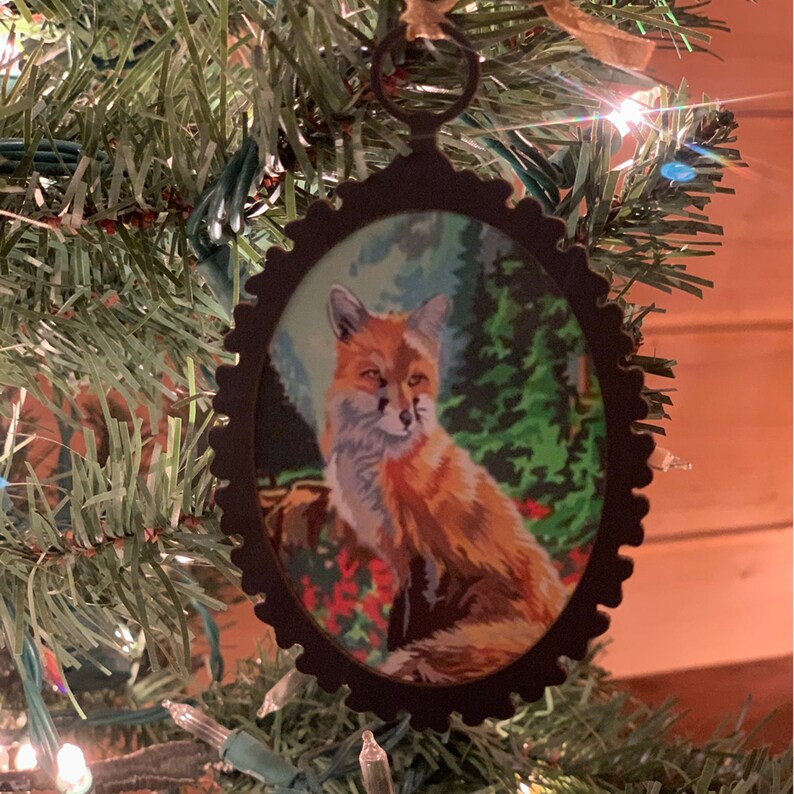 May include: A brown metal Christmas ornament with a fox illustration. The fox is orange and white with a black nose and eyes. The ornament is surrounded by a green forest scene.