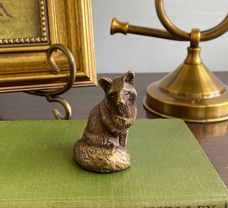 May include: A small, gold-colored metal figurine of a fox sitting on a green book. The fox is detailed with fur and has a curled tail.