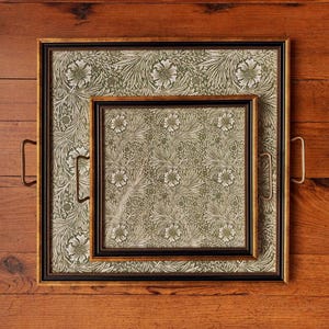 May include: Two decorative trays with floral patterned inserts. The larger tray has a gold frame with brass handles, and the smaller tray has a black frame. The floral pattern features white flowers and green leaves on a cream background.