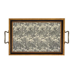 May include: Rectangular serving tray with a black and gold frame and brass handles. The tray's interior features a cream-colored background with a repeating pattern of black and gray floral and leaf designs. A decorative piece for serving.