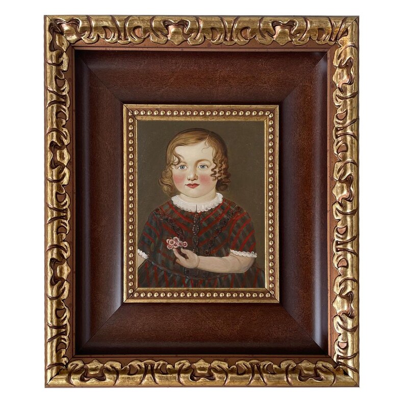 May include: A framed portrait of a young child. The portrait is set within a dark brown frame with a gold ornate outer frame. The child is wearing a red and black patterned dress and holding a small flower.