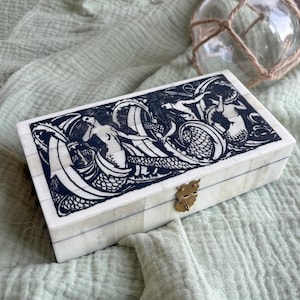 May include: A rectangular box with a white tiled surface featuring a black ink illustration of mermaids swimming among waves on the lid. The box has a brass-coloured metal latch on the front and is constructed from small, joined rectangular pieces.