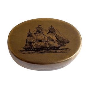 May include: A circular, wooden trinket box with a gold finish. The lid displays a detailed black illustration of a tall sailing ship. The box has a slightly raised edge.