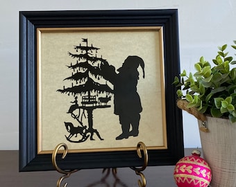 May include: A black silhouette of Santa Claus decorating a Christmas tree with ornaments and a toy train. The silhouette is framed in a black frame with a gold inner border and is displayed on a gold metal stand.