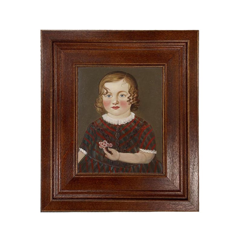 May include: A framed portrait of a young child. The child has curly blonde hair, rosy cheeks, and is wearing a red, black, and white patterned dress. The child is holding a small flower. The frame is a dark brown color.
