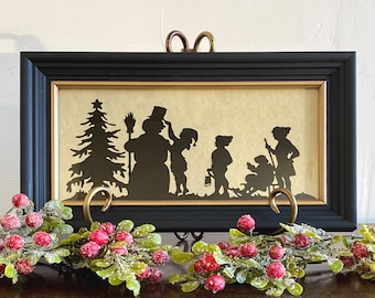 May include: Black silhouette of a snowman and children in a winter scene framed in a black frame with a gold mat. The image is on a dark wood surface with a green and red garland.