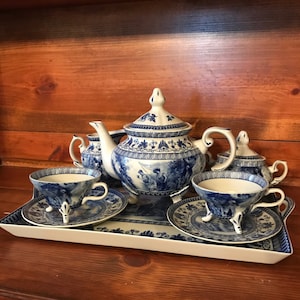 May include: A blue and white porcelain tea set with a teapot, creamer, sugar bowl, and two teacups and saucers. The set features a floral design and a scene of people fishing. The set is on a white tray with a blue and white floral design.