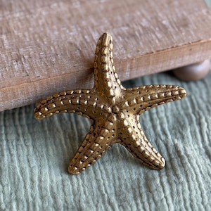 Op de afbeelding: Een gouden zeestersculptuur met gedetailleerde textuur. De vijfvingerige zeester wordt getoond op een licht houten oppervlak tegen een zachte, lichtgroene stoffen achtergrond. Dit decoratieve stuk roept een kust- of nautisch thema op.